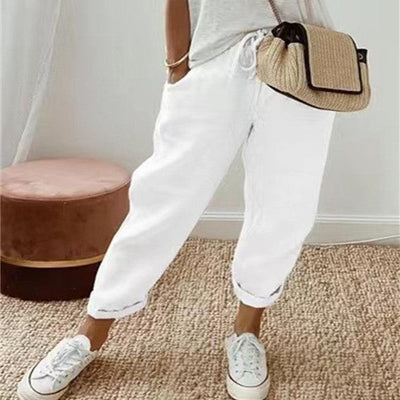 Femme portant un pantalon décontracté uni blanc avec sneakers blanches et sac bandoulière tissé, ambiance intérieure cosy.