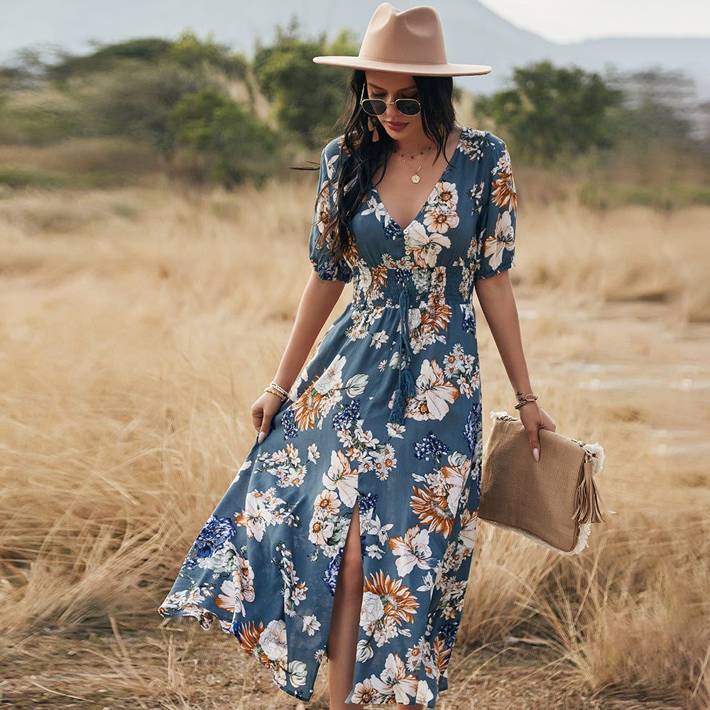 Femme portant une robe d'été fleurie bleue avec motifs floraux, chapeau beige et lunettes de soleil dans un champ sec.