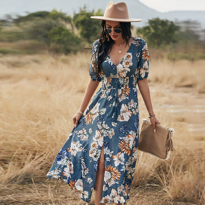 Femme portant une robe d'été fleurie bleue avec motifs floraux, chapeau beige et lunettes de soleil dans un champ sec.
