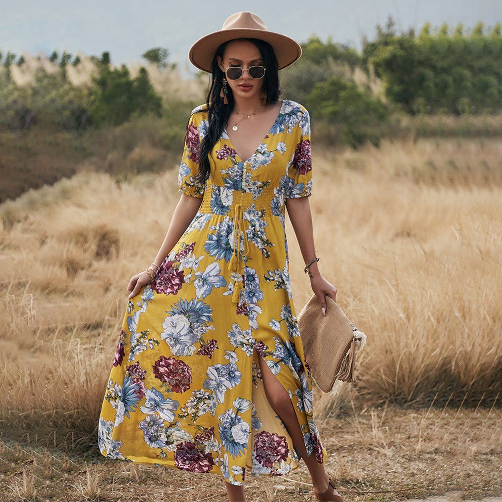 Femme portant une robe d'été fleurie jaune élégante avec motifs floraux et fente, accessoirisée d'un chapeau beige et lunettes de soleil en plein air champêtre.