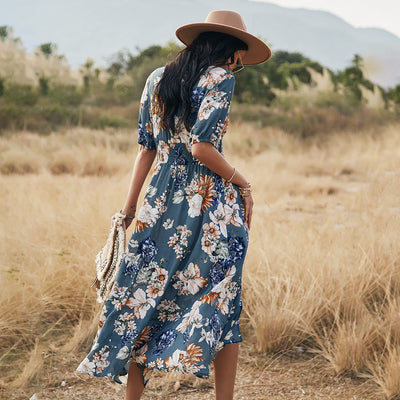 Femme portant une robe d'été fleurie bleue fluide avec motifs floraux, chapeau beige, dans un champ sec avec montagnes en arrière-plan.